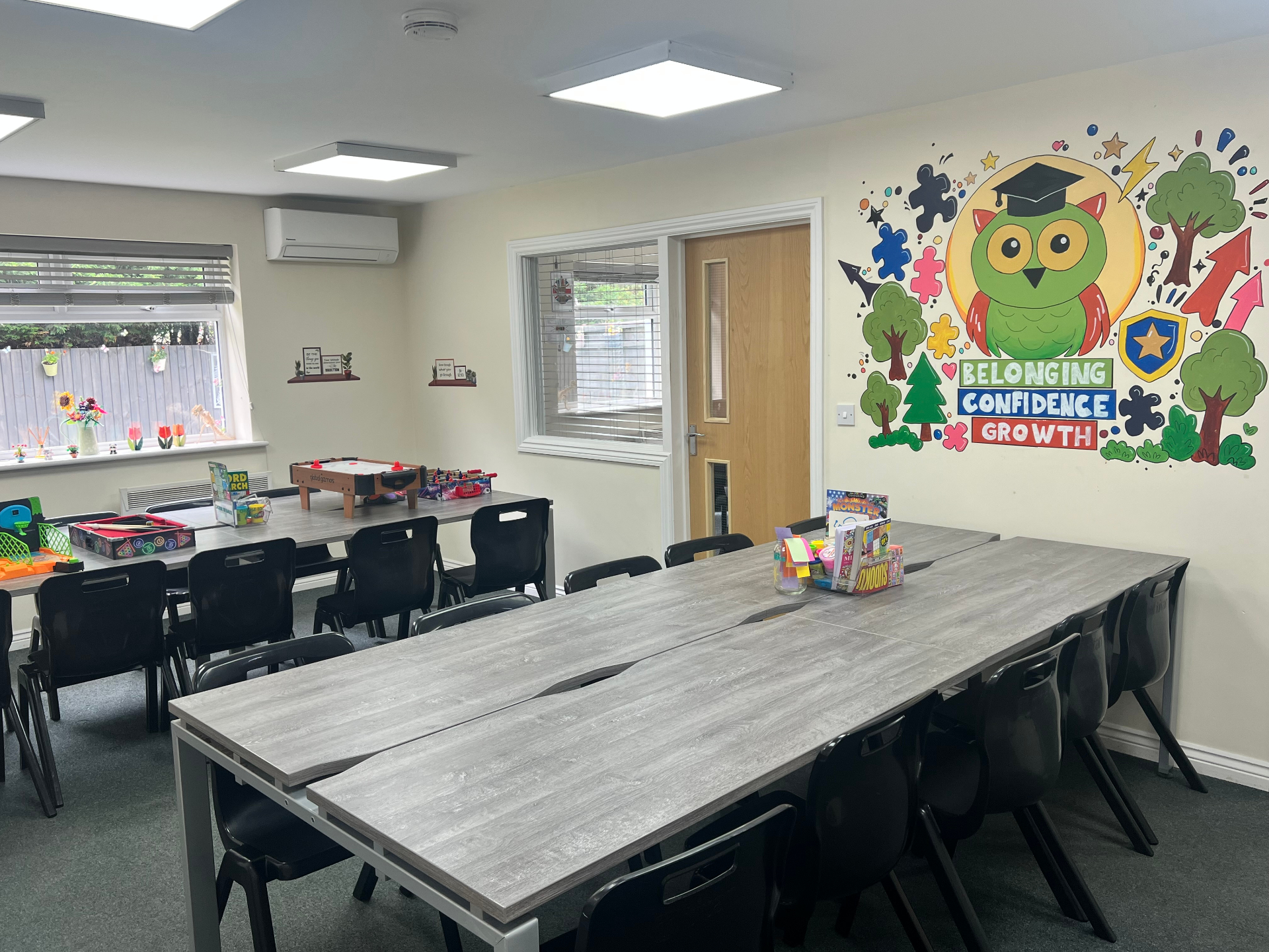 A bright classroom with shared tables, black chairs, games and a colourful owl mural celebrating belonging, confidence and growth