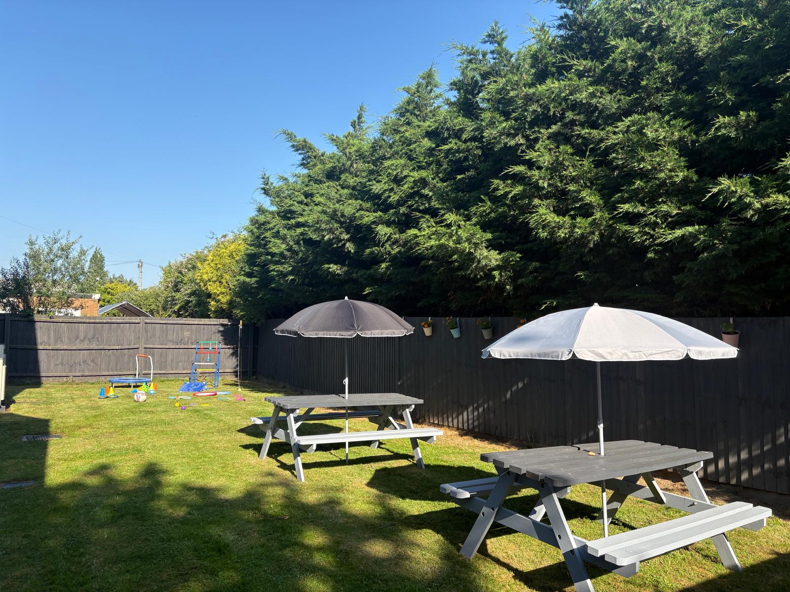 A sunny outdoor garden with picnic benches, parasols, play equipment and tall trees along the fence