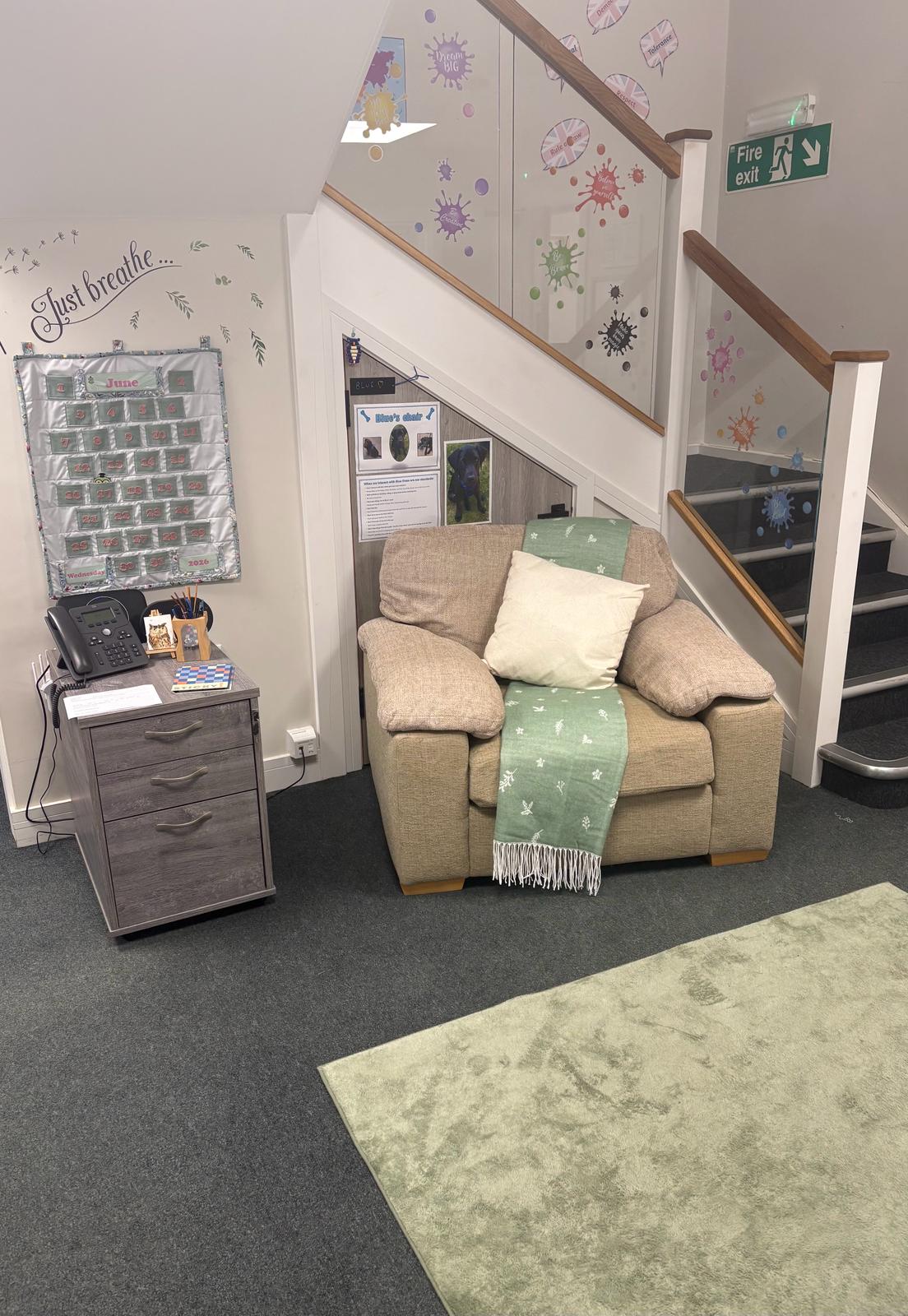 A cosy corner under the stairs with a soft sofa, 'Just breathe' wall art, a June calendar and gentle decor