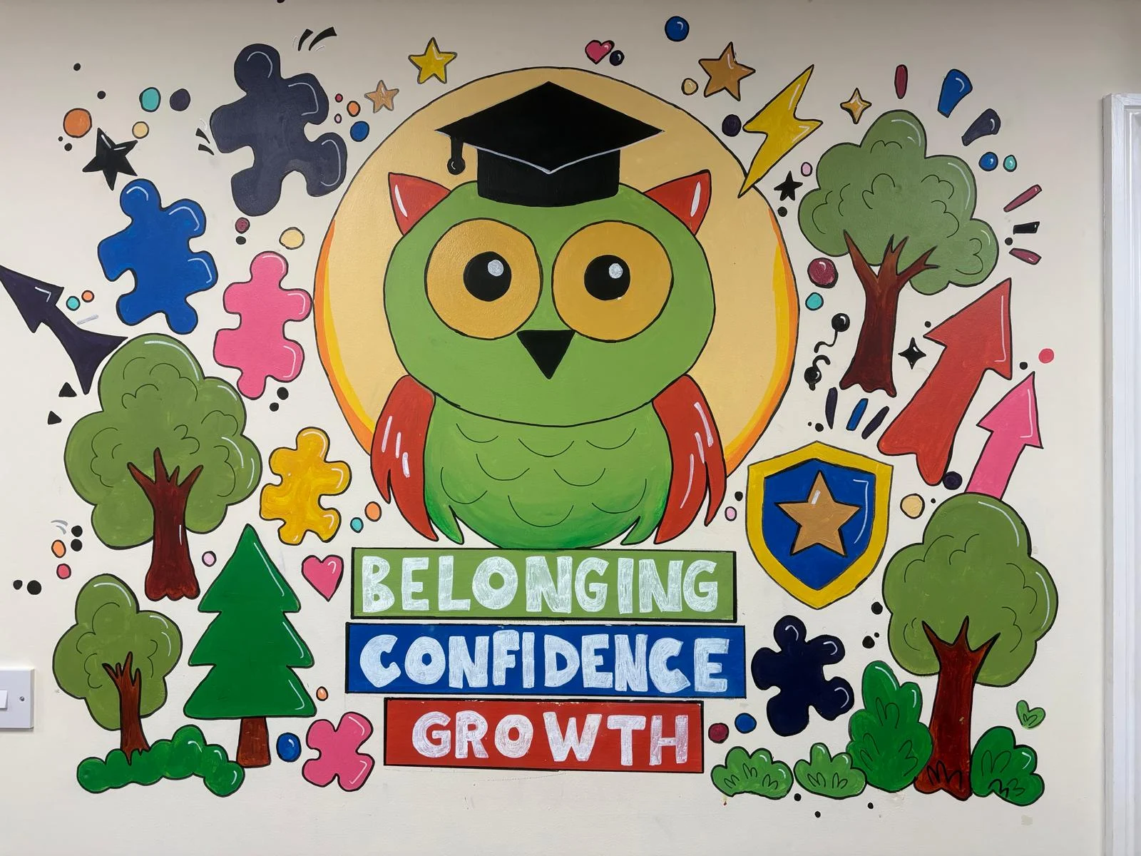 A colourful school mural featuring a wise owl wearing a graduation cap, surrounded by trees, puzzle pieces, stars and the words Belonging, Confidence, Growth