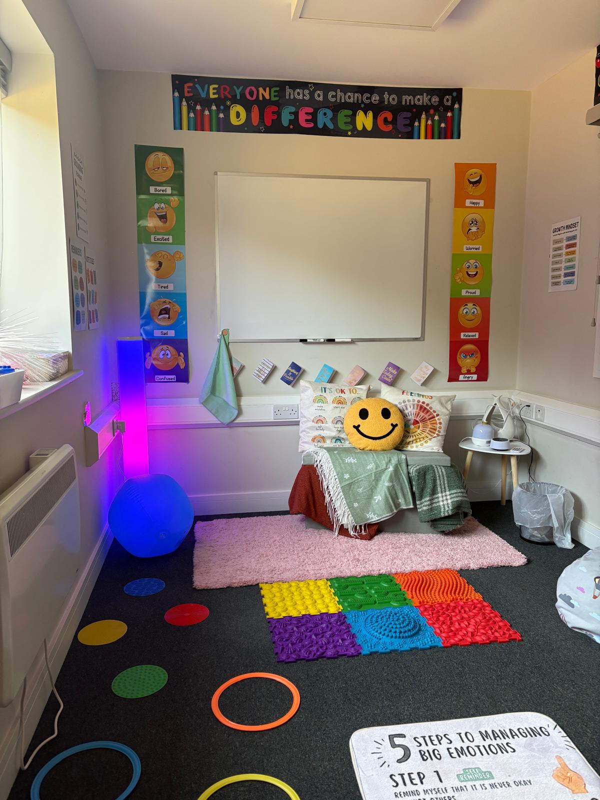 A sensory calm room with soft seating, emotion posters, colourful textured mats, a smiley cushion and gentle coloured lighting